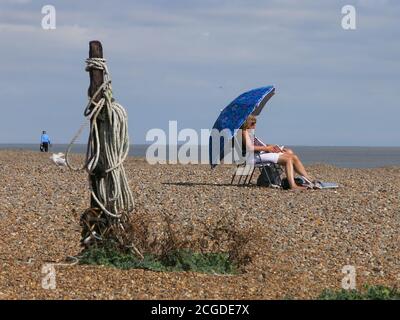 Eine Frau sitzt und liest unter einem großen blauen Sonnenschirm am Kiesstrand von Aldeburgh. Spiralseil und Treibholz tragen zur Seeszene bei. Stockfoto