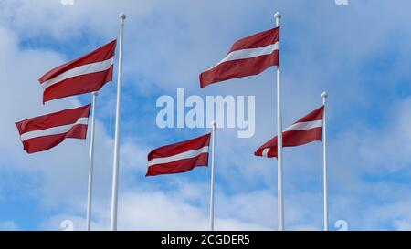 Lettland Flaggen winken im Wind gegen einen blauen Himmel. Flagge Lettlands auf blauem und wolkenverhangenem Himmel Hintergrund Fliegen im starken Wind. Lettische Nationalflagge. Stockfoto