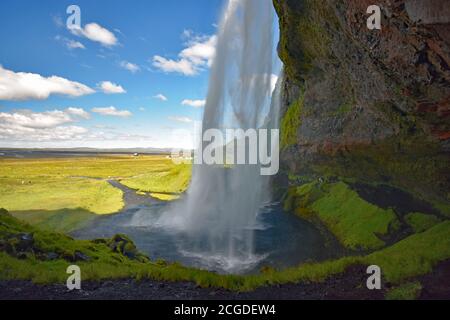 Ein Blick von der Seite des Seljalandsfoss Wasserfalls an einem hellen sonnigen Tag in Südisland. Hinter den Wasserfällen kann man die Höhle und den Wanderweg sehen. Stockfoto