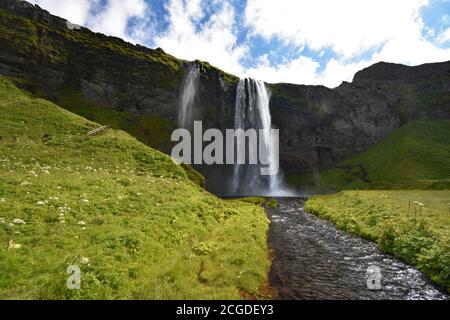 Wasser von Eyjafjallajökull, wie es über die Klippe schafft Seljalandsfoss in Süd-Island fließt. Ein Fluss fließt durch Felder von hellgrünem Gras. Stockfoto