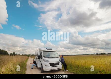 Eine Frau schaut in den Himmel, ein Mann sitzt auf der Stufe eines Autos, an einem Wohnmobil auf einer Landstraße, inmitten von Feldern. Sommer Sonnentag. Paar nach 50 Jahren, Urlaubskonzept. Hochwertige Fotos Stockfoto