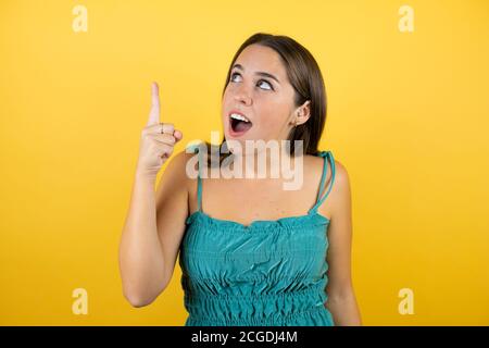 Junge schöne Frau über isolierten gelben Hintergrund überrascht, zeigt und schaut nach oben. Stockfoto