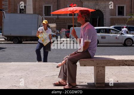 Rom, Italien - 28. Juni 2010: Ein Straßenverkäufer von Regenschirmen, bleibt auf einer Bank sitzen, neben einer Frau, die als Reiseleiter in Rom arbeitet. Stockfoto