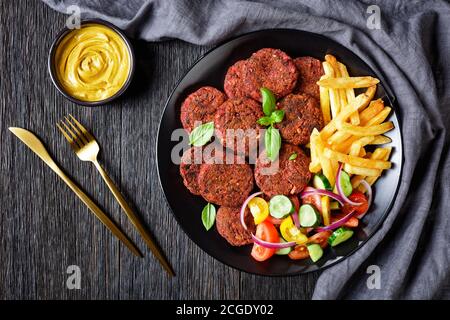 Vegane Rote-Beete-Kuchen aus Pilzen, schwarze Bohnen serviert mit pommes Frites und frischer Gemüsesalat auf einem schwarzen Teller mit Hefesauce auf einem da Stockfoto