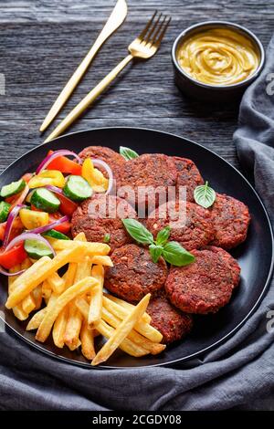 Vegane Rote-Beete-Patties aus Pilzen, schwarze Bohnen mit pommes Frites und frischer Gemüsesalat auf einem schwarzen Teller mit veganer Käsesauce auf einem dunklen Stockfoto