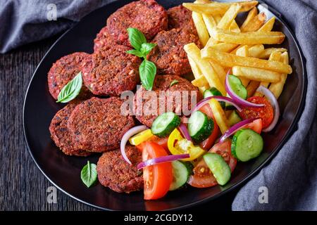 Vegane Mahlzeit: Rote Beete Kuchen mit Pilzen und schwarzen Bohnen serviert mit pommes Frites und Gemüsesalat mit Tomaten, Gurken und roten Zwiebeln auf einem schwarzen Teller Stockfoto