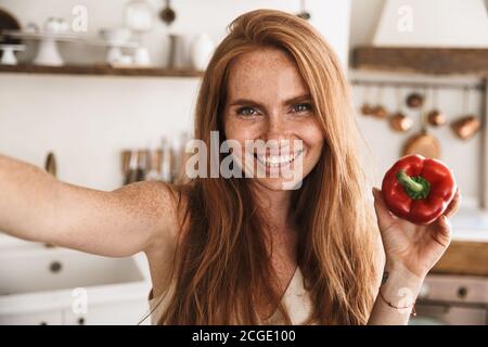 Bild von glücklichen Ingwer Frau macht Selfie Foto auf Handy Telefon mit Paprika beim Kochen in der gemütlichen Küche Stockfoto