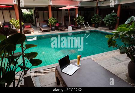 Ein weißer Laptop, Smartphone und Mango Smoothie auf einer Sonnenliege vor dem Pool Hintergrund. Ein Start in den neuen Tag. Freiberufliches Geschäftskonzept. Flexible RE Stockfoto