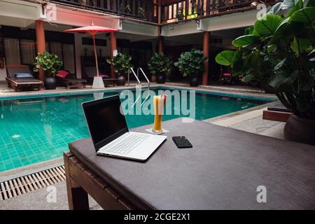 Ein weißer Laptop, Smartphone und Mango Smoothie auf einer Sonnenliege vor dem Pool Hintergrund. Ein Start in den neuen Tag. Freiberufliches Geschäftskonzept. Flexible RE Stockfoto