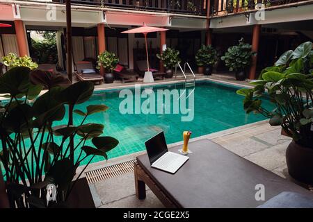 Ein weißer Laptop, Smartphone und Mango Smoothie auf einer Sonnenliege vor dem Pool Hintergrund. Ein Start in den neuen Tag. Freiberufliches Geschäftskonzept. Flexible RE Stockfoto