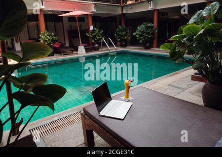 Ein weißer Laptop, Smartphone und Mango Smoothie auf einer Sonnenliege vor dem Pool Hintergrund. Ein Start in den neuen Tag. Freiberufliches Geschäftskonzept. Flexible RE Stockfoto