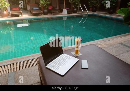 Ein weißer Laptop, Smartphone und Mango Smoothie auf einer Sonnenliege vor dem Pool Hintergrund. Ein Start in den neuen Tag. Freiberufliches Geschäftskonzept. Flexible RE Stockfoto