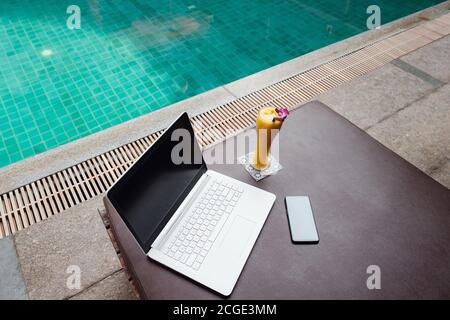 Ein weißer Laptop, Smartphone und Mango Smoothie auf einer Sonnenliege vor dem Pool Hintergrund. Ein Start in den neuen Tag. Freiberufliches Geschäftskonzept. Flexible RE Stockfoto