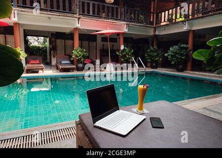 Ein weißer Laptop, Smartphone und Mango Smoothie auf einer Sonnenliege vor dem Pool Hintergrund. Ein Start in den neuen Tag. Freiberufliches Geschäftskonzept. Flexible RE Stockfoto