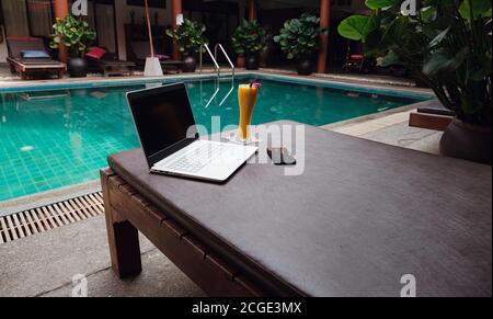 Ein weißer Laptop, Smartphone und Mango Smoothie auf einer Sonnenliege vor dem Pool Hintergrund. Ein Start in den neuen Tag. Freiberufliches Geschäftskonzept. Flexible RE Stockfoto