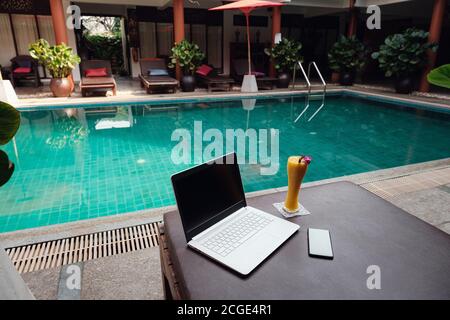 Ein weißer Laptop, Smartphone und Mango Smoothie auf einer Sonnenliege vor dem Pool Hintergrund. Ein Start in den neuen Tag. Freiberufliches Geschäftskonzept. Flexible RE Stockfoto