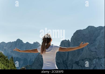 Blick von hinten auf eine junge Wanderin, die mit offenen Armen das Leben genießt und den Blick auf die Alpen genießt. Stockfoto