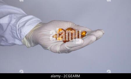 Eine Hand in einer weißen Uniform und ein Gummihandschuh hält gelbe Tabletten - Fischöl Kapseln. Stockfoto