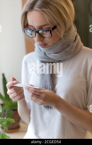 Junge Frau in Brillen in Schal gewickelt fühlt sich kalt Symptom und Blick auf Thermometer. Virus, Grippesaison Stockfoto