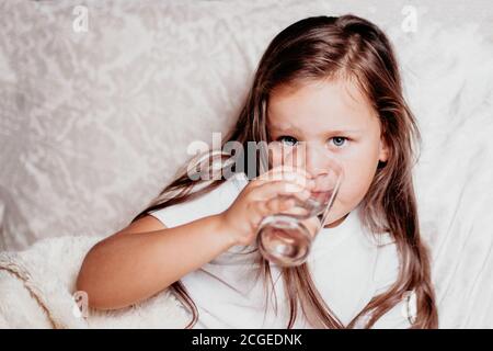 Ein krankes Kind, ein Mädchen, das auf einem Bett mit einem weißen Kissen sitzt und Wasser aus einem Glas trinkt Stockfoto