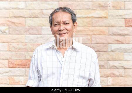 Porträt eines lächelnden asiatischen älteren Mannes, der mit selbstbewusster Art und Weise vor Backsteinmauer steht. Nahaufnahme Gesicht von älteren Lächeln mit grauen Haaren Stockfoto