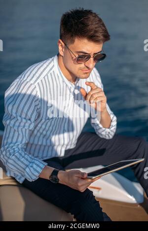 Leerzeichen für Text. Schöner junger Mann auf Bürokleidung entspannen und arbeiten auf digitalen Tablet auf Motorboot Bogen, über Meer Hintergrund Stockfoto