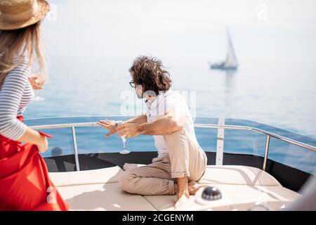 Europäische attraktive braunhaarige Mann in spectaces und blonde Frau mit Strohhut und abisolierten Bluse, umarmen, Chillen mit Freunden am Segeln Stockfoto
