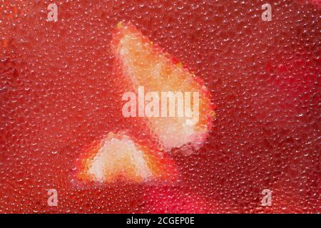 Rote Frucht Hintergrund, erfrischend in der Hitze. Wassertropfen auf dem Glas eines Behälters mit hausgemachter Beerenlimonade. Erdbeeren hinter g Stockfoto