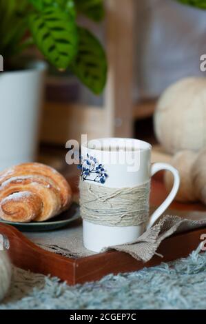 Weißer Tee Tasse mit gestrickten Fäden und Croissant in Holztablett auf grünem Pflanzenhintergrund. Gemütliches Frühstück Stockfoto