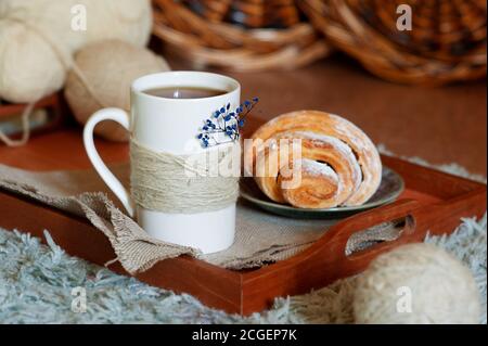 Weißer Tee Tasse mit gestrickten Fäden und akroissant in Holztablett auf kuscheligem Teppich, Korbkörbe und Kugeln aus Wollfäden auf Hintergrund Stockfoto