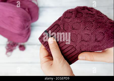 Hände halten kastanienbraunen Strickpullover mit Muster und Kugeln aus Wollfäden daneben auf hellem Holzhintergrund. Strickstunden online Stockfoto