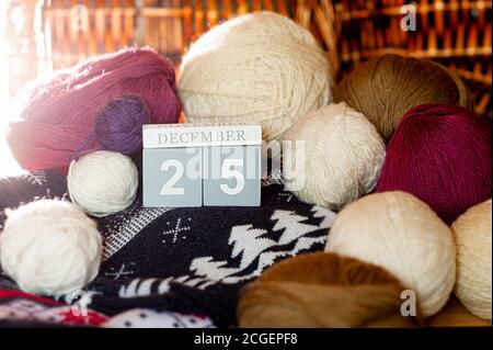 Holzkalender mit dem Datum Dezember 25, Kugeln aus bunten Wollfäden zum Stricken und warmer Pullover mit Wintermuster, festliche Weihnachten c Stockfoto