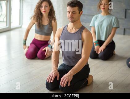 Positive sportliche männliche Instruktor, die Yoga-Klasse in Fitness-Studio mit grauem Hintergrund Durchführung Asana mit fit Frauen Schüler auf Hintergrund. Aktiv Stockfoto