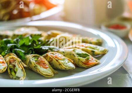 Brötchen aus dünnen Pfannkuchen mit Lachs und Frischkäse auf einem weißen Teller mit frischer Petersilie grün. Nahaufnahme, selektiver Fokus Stockfoto