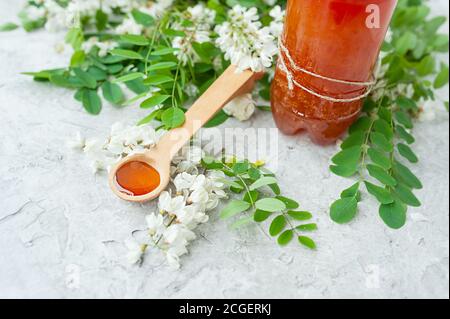 Akazienhonig in einem Holzlöffel mit üppig blühenden weißen Akazienblüten und Flasche Honig auf dem Hintergrund, analoge Medizin, nützliche Eigenschaften der Pflanzen Stockfoto