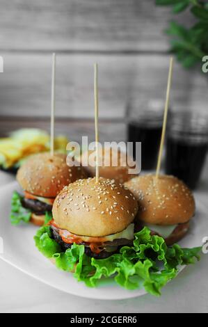 Junk Food, hausgemachte Hamburger aus Brötchen mit Sesam, Schnitzel, Sauce und frischem Salat auf langen Holzspießen, zwei Gläser Schwarzgetränk auf einem Woo Stockfoto