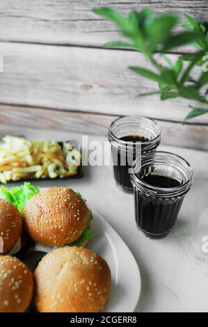 Junk Food, zwei Gläser Schwarzgetränk und hausgemachte Hamburger aus Brötchen mit Sesam, Cutlet, Sauce und frischem Salat auf einem Holztisch Stockfoto