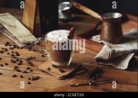 Glas transparent Kaffee Tasse mit Doppelwänden mit Kaffee gegossen In Schichten mit Zimt und Milchschaum auf rauem Holz Tisch und Kaffee verstreut bea Stockfoto