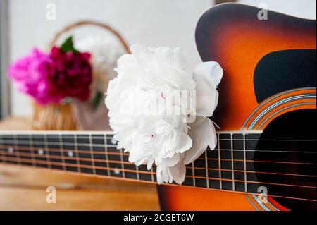 Weiße frische üppige Pfingstrose auf dem schwarzen Griffbrett eines Sechs Saiten Holzgitarre und verschwommene bunte Pfingstrosen in A Korbkorb im Hintergrund Stockfoto