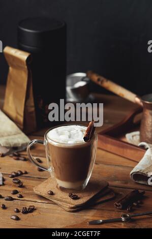 Glas transparent Kaffee Tasse mit Doppelwänden mit Kaffee gegossen In Schichten mit Zimt und Milchschaum auf rauem Holz Tisch und Kaffee verstreut bea Stockfoto