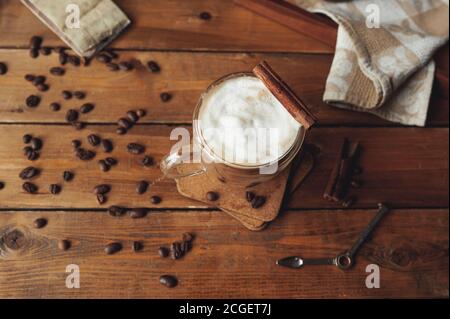 Glas transparent Kaffee Tasse mit Doppelwänden mit Kaffee gegossen In Schichten mit Zimt und Milchschaum auf rauem Holz Tisch und Kaffee verstreut bea Stockfoto