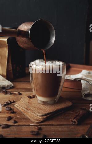 Türkische Kaffeekanne gießt ein Getränk in Glas transparenten Kaffee Tasse mit doppelten Wänden mit Kaffee gegossen in Schichten mit Zimt und Milchschaum auf rauem w Stockfoto