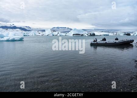 Ein Tierkreisboot, das in den flachen Gewässern auf der Seeseite der Jökulsárlón Glacier Lagoon, Südisland, gebunden ist. Eisberge schweben dahinter Stockfoto