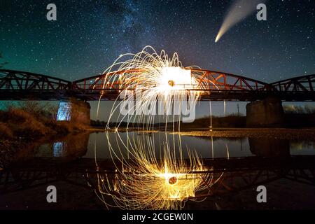 Spinnende Stahlwolle im abstrakten Kreis, Feuerwerksschauer von leuchtend gelben Funken auf einer langen Brücke, die sich im Flusswasser unter dem dunklen Sternenhimmel der Nacht spiegeln. Stockfoto