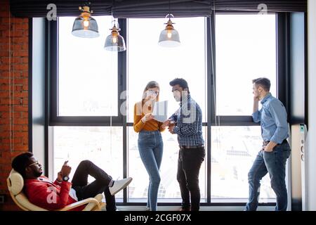 Multiethnische Gruppe von Studenten chillen in der Universitätshalle während der Pause, stehen am Fenster reden oder kommunizieren in sozialen Medien über Laptop Stockfoto