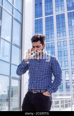 Indischer ernster Mann in legerem Tuch mit Bart, der mit seinem Telefon in der Nähe seines Ohrs vor einem Bürogebäude steht. Stockfoto