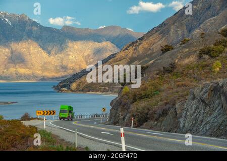 Das grüne Auto läuft den Hügel hinunter in eine kurvige Straße. Sehen Sie das Meer und die Berge am Abend und den blauen Himmel am See Hawea, Wanaka Otago, New Stockfoto