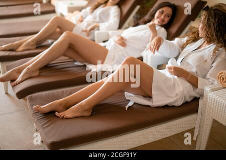 Fröhliche junge Freundinnen beginnen Geburtstagsfeier im Spa-Center, liegen in weißen Bademänteln auf Liegestühlen nach einer Schaummassage, Demonstration perf Stockfoto