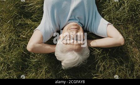 Ältere Frau, Rentnerin auf dem Gras liegend mit den Händen unter dem Kopf. Oben Nahaufnahme. Hochwertige Fotos Stockfoto