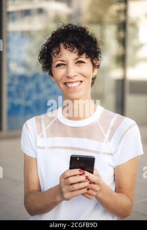 Porträt von lächelnden reifen Frau mit kurzen schwarzen Haaren halten Handy in der Stadt Stockfoto